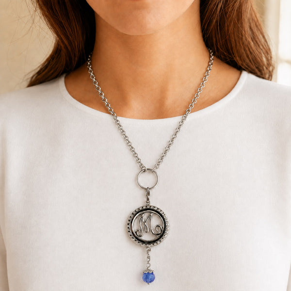 A close-up of a woman wearing a Floating Charm Locket necklace featuring a cursive monogram and a dangling blue bead, paired with a white top.