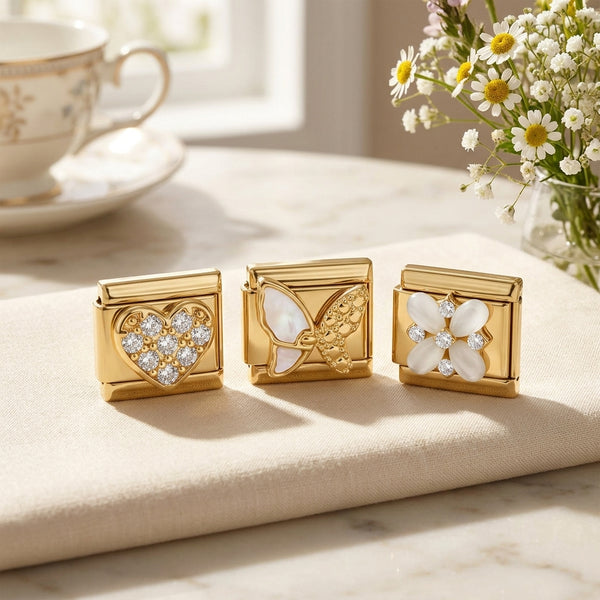 Three gold9mm Italian charms with floral, butterfly and crystal heart design on a light surface with a cup and flowers in the background.
