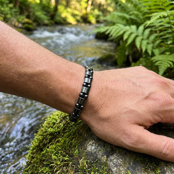 Person's wrist wearing a black beaded hematite bracelet with a natural background of water and greenery.