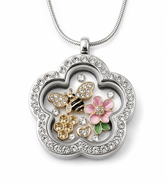 A silver floating charm locket necklace in a flower-shape with clear crystals. The pendant is decorated with a silver rhinestone border, a gold and black bee, a pink flower with green leaves, and small heart charms, all suspended inside.
