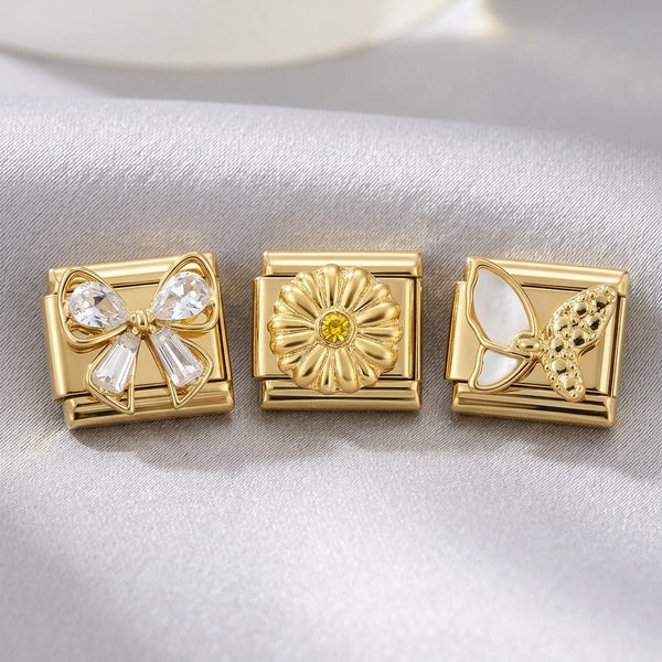 Three gold 9mm Italian charms in crystal bow, citrine flower and white enamel butterfly designs on a light fabric background