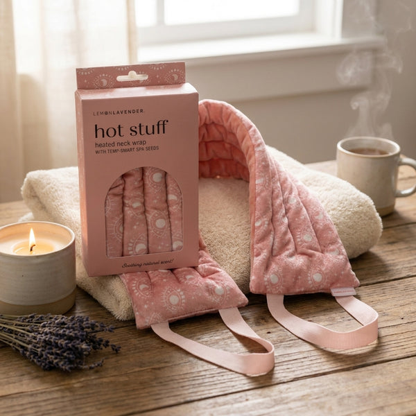 Pink heated neck wrap with packaging on a wooden surface with a candle and cup of coffee in the background.