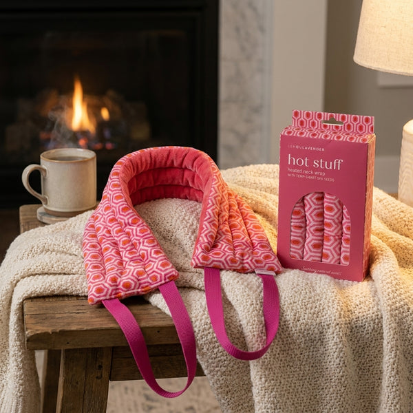 Hot Stuff by Lemon Lavender pink geometric heated neck wrap on a wooden stool and blanket in front of a fireplace.