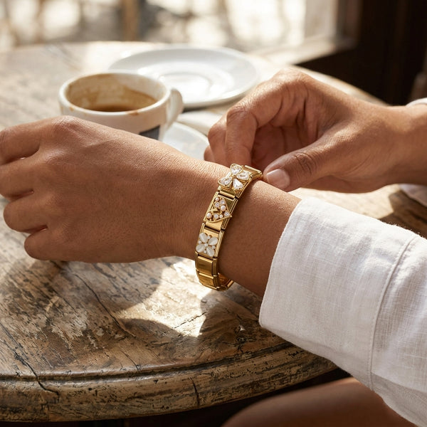 Person adjusting a gold 9mm Italian charm bracelet on a wooden table with a cup of coffee in the background