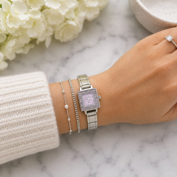 Silver-toned square 9mm Italian charm watch with a light purple dial and crystal-encrusted bezel, shown on a woman's wrist alongside two delicate silver bracelets, resting on a white marble surface with white flowers in the background.