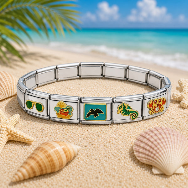 A silver Italian charm bracelet with colorful enamel tropical-themed charms, displayed on a sandy beach with seashells and a blurred ocean background.